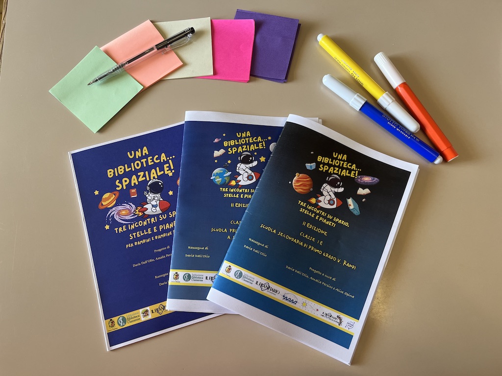 Tre libricini, uno per ogni edizione di Una biblioteca spaziale. Vicino ai libretti ci sono dei post-it colorati, pennarelli e una penna.